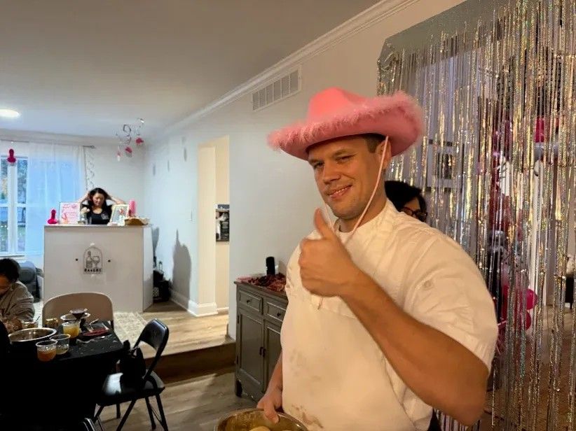 Man in a pink fuzzy cowboy hat giving a thumbs-up at a living-room party, holding a mixing bowl in front of a silver fringe backdrop with pink decorations and a snack table.