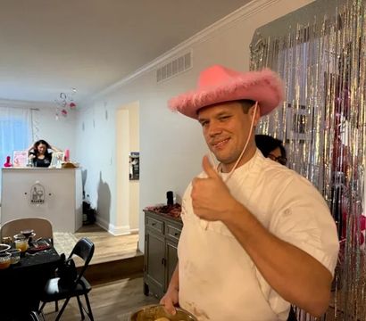 Man in a pink fuzzy cowboy hat giving a thumbs-up at a living-room party, holding a mixing bowl in front of a silver fringe backdrop with pink decorations and a snack table.