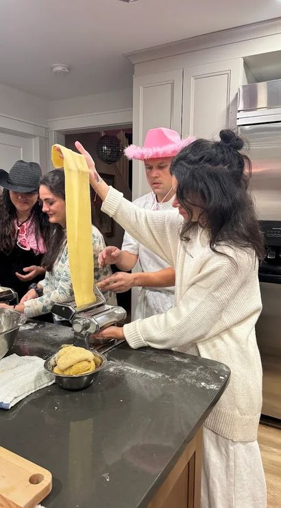 People in a home kitchen making homemade pasta — a long sheet of dough fed through a pasta machine on a flour-dusted island with bowls of dough and playful hats.