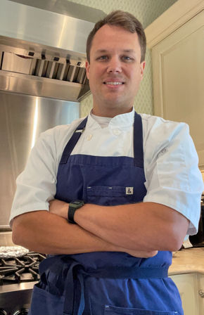 Smiling chef wearing a white coat and navy blue apron with arms crossed in a kitchen beside a stainless-steel stove and range hood