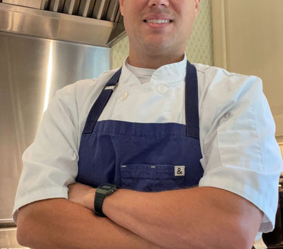 Smiling chef wearing a white coat and navy blue apron with arms crossed in a kitchen beside a stainless-steel stove and range hood