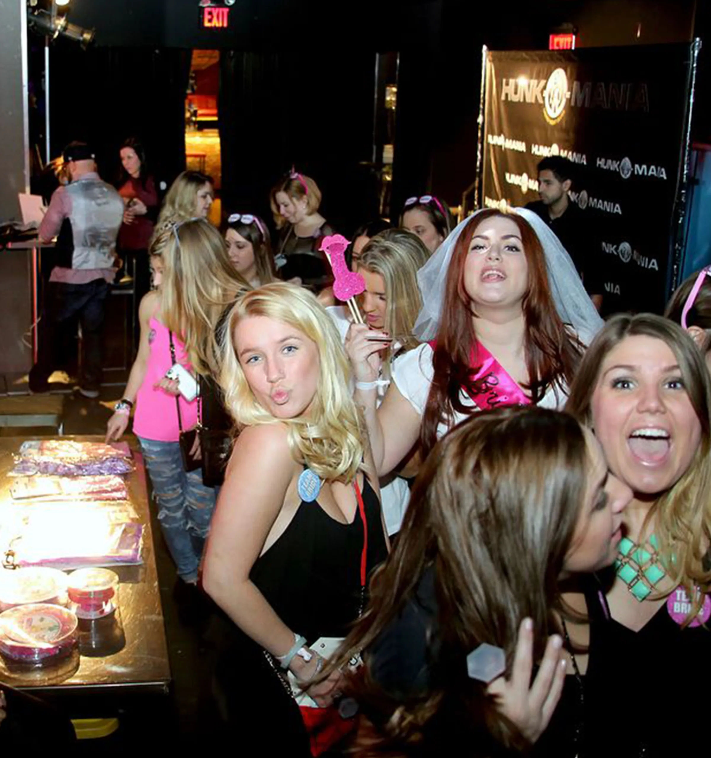Group of women at a lively nightclub bachelorette party, bride-to-be wearing a veil and pink sash holding a glittery prop while friends pose, laugh, and grab party favors.