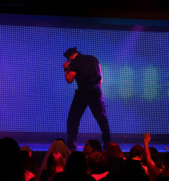 Spotlighted male performer in dark costume and hat on a nightclub stage with a purple-blue LED wall backdrop, dancing while a silhouetted crowd reaches up