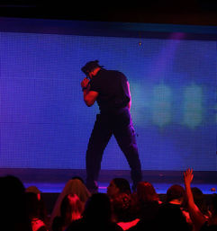 Male dancer in a police costume striking a pose on a colorful LED nightclub stage with a cheering audience