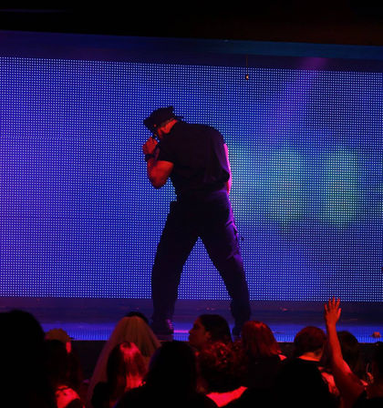 Male dancer in a police costume striking a pose on a colorful LED nightclub stage with a cheering audience