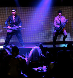 Two dancers in trench coats and fedoras performing with umbrellas on a neon-lit nightclub stage, purple spotlight and LED backdrop, silhouetted audience seated at tables in the foreground.