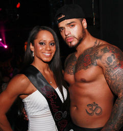 Smiling woman wearing a "Bride to Be" sash poses with a shirtless, heavily tattooed man in a dim nightclub — bachelorette party vibe with colorful party lighting.