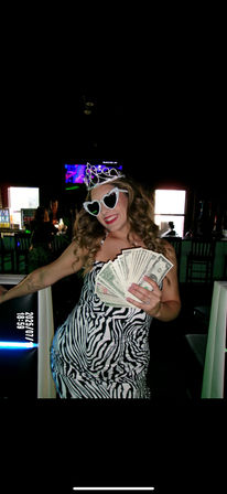 Woman in zebra-print dress wearing a tiara and rhinestone heart-shaped sunglasses, smiling and holding a fan of cash in a dimly lit bar with neon lighting — celebratory nightlife scene.