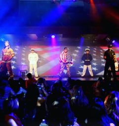 Patriotic stage show with performers in firefighter, sailor, soldier draped in a U.S. flag, marine and police uniforms under colorful stage lights, live audience watching in the foreground.