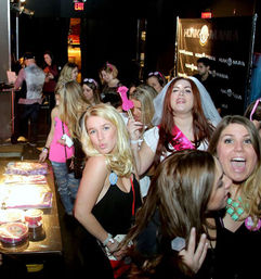 Bachelorette party at a lively nightclub — group of women posing and laughing, bride-to-be wearing a veil and sash, friends holding pink party props and taking photos.