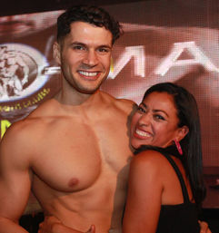 Smiling shirtless muscular man posing with a grinning woman hugging him in front of a lit LED event stage backdrop