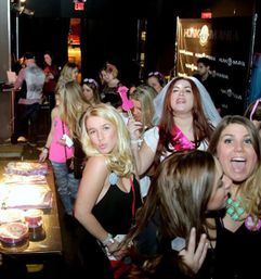 Bachelorette party in a nightclub: bride in a veil and pink sash holding a glitter flamingo prop while a group of friends laugh, pose, and gather near a table with party treats.