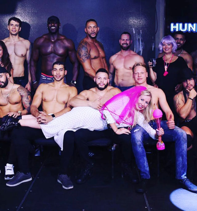 Playful bachelorette party photo: bride-to-be in a pink veil lying across the laps of shirtless male dancers on a nightclub stage, holding a pink novelty drink.