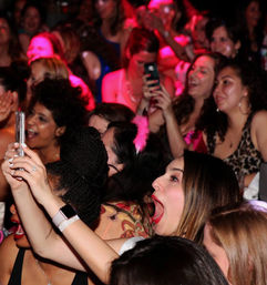 Excited group of women packed in a nightclub, cheering and taking photos with smartphones under pink party lights