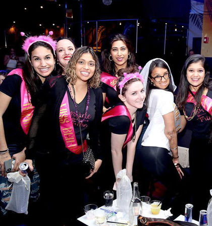 Smiling women in pink bachelorette sashes, tiaras and a veil posing at a lively nightclub table with cocktails — girls' night out.
