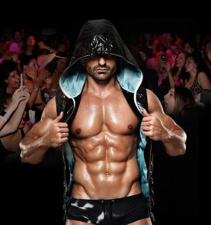 Muscular male performer in a glossy hooded vest pulling it open to reveal a chiseled, oiled six-pack while an excited nightclub crowd of women cheers and takes photos.