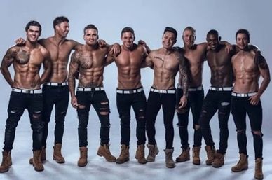 Eight shirtless male fitness models in ripped black jeans and tan work boots, standing arm-in-arm and showing toned abs and tattoos in a studio photoshoot.