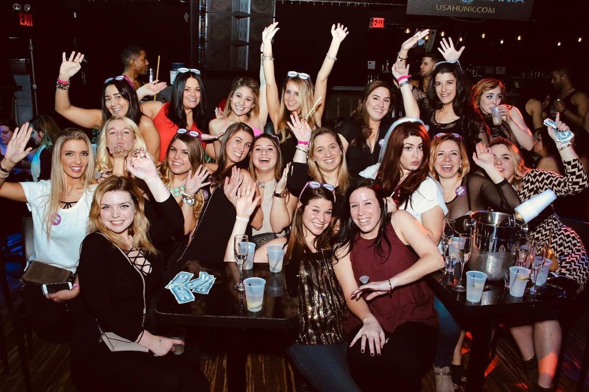 Cheerful group of women partying at a lively nightclub table for a bachelorette/girls' night out, smiling, raising hands and holding drinks.