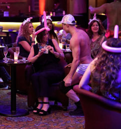 Nightclub lounge scene: women wearing balloon hats and sipping champagne watch a shirtless male dancer in a silver cap and shorts as one guest tips him with cash.