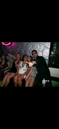 Neon-lit nightclub booth with a bachelorette party: smiling women (one wearing a ‘Bride to Be’ sash) with dollar bills tucked into outfits, a man pointing a microphone/prop at the camera against a textured silver wall.