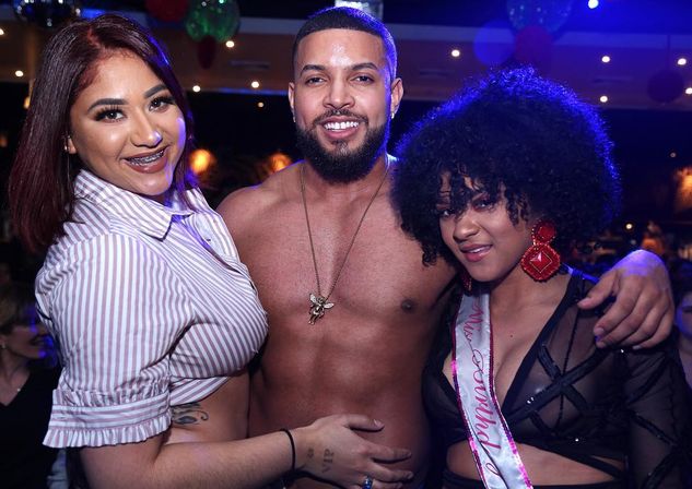 Three friends posing at a lively nightclub party: a shirtless smiling man with a pendant in the center, flanked by a woman in a striped crop top with braces and a woman in a black sheer outfit wearing a sash and red statement earrings, colorful party lights in the background.