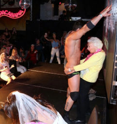 Nightclub stage scene at a bachelorette party: a scantily clad male performer poses while an older woman in a yellow sweater embraces him, veiled guests cheer and disco balls glitter overhead.