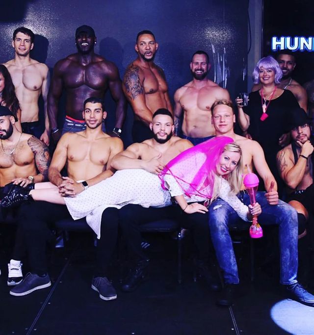 Group photo of shirtless male dancers on a nightclub stage with a smiling woman in a pink veil holding a novelty drink, posing in a playful bachelorette-party vibe.