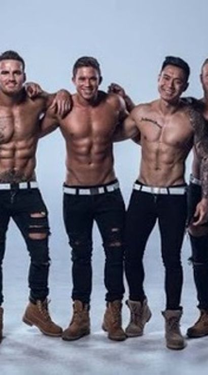 Eight shirtless muscular male fitness models in black jeans and tan work boots posing arm-in-arm and smiling in a light studio photoshoot