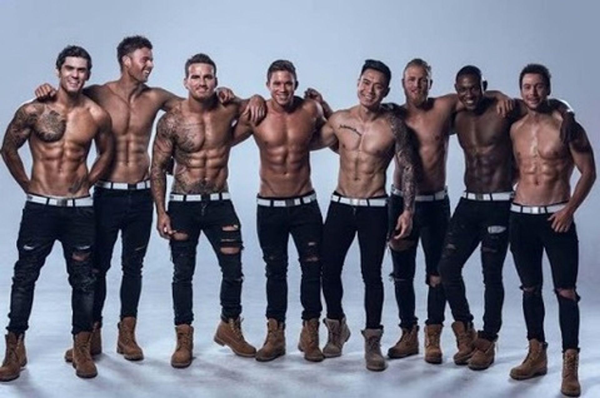 Eight shirtless muscular male fitness models in black jeans and tan work boots posing arm-in-arm and smiling in a light studio photoshoot