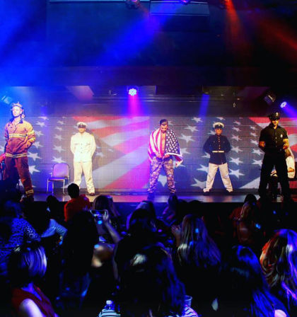 Patriotic stage show with five male performers in firefighter, naval, camouflage (wrapped in a U.S. flag), marine and police uniforms in front of a large American flag backdrop and colorful lights, audience watching.