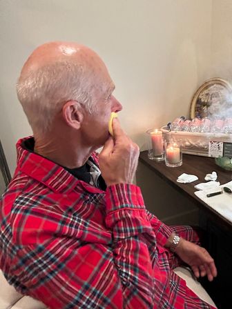Bald man in a red plaid shirt dabbing his face with a yellow sponge at a cozy home vanity table with lit candles, bottled water, a mirror and cotton pads.