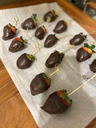 Glossy chocolate-covered strawberries skewered with bamboo picks, arranged on parchment-lined wooden board — party-ready dessert