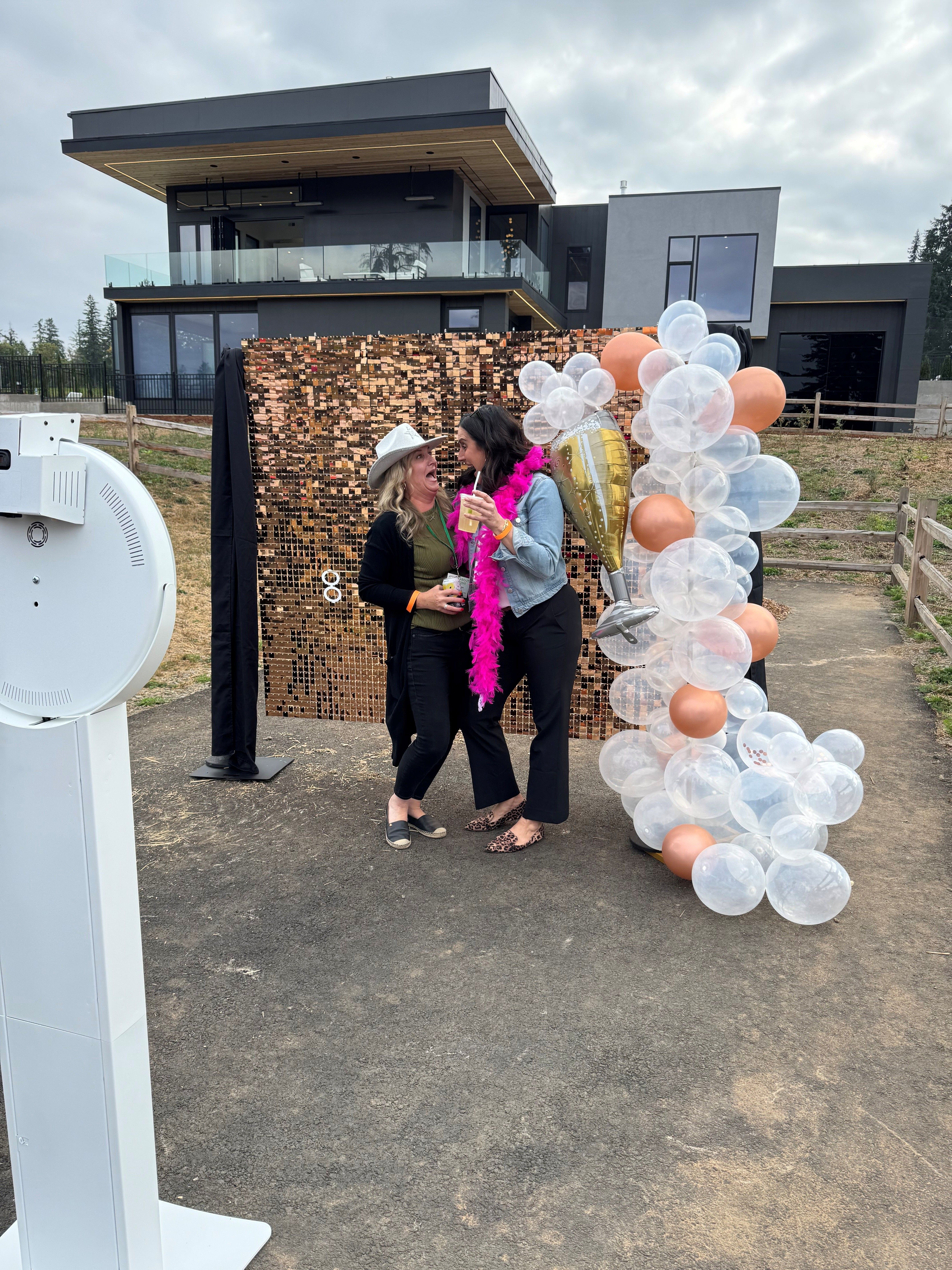 Two people laughing at an outdoor photo booth with a rose-gold sequin backdrop and clear-and-rose-gold balloon garland, one wearing a pink feather boa and holding drinks in front of a modern multi-level house at a casual backyard party