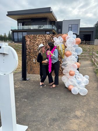 Two people laughing at an outdoor photo booth with a rose-gold sequin backdrop and clear-and-rose-gold balloon garland, one wearing a pink feather boa and holding drinks in front of a modern multi-level house at a casual backyard party