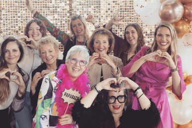 Smiling group of women posing in a glitter photo-booth backdrop with rose-gold and white balloons, making heart hand gestures and wearing colorful outfits and playful props like a pink feather boa and a 'PAPARAZZI' sign.