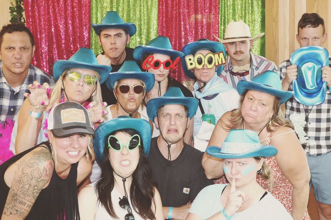 Party photo booth group wearing turquoise cowboy hats and silly props (oversized “COOL”/“BOOM” glasses, alien shades) making funny faces against a pink and green sequin backdrop.