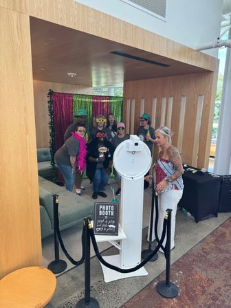 Group of seven adults in colorful hats, sunglasses and a pink feather boa posing at a white photo-booth kiosk in a modern indoor lobby with green and magenta sequin backdrop and velvet stanchions.