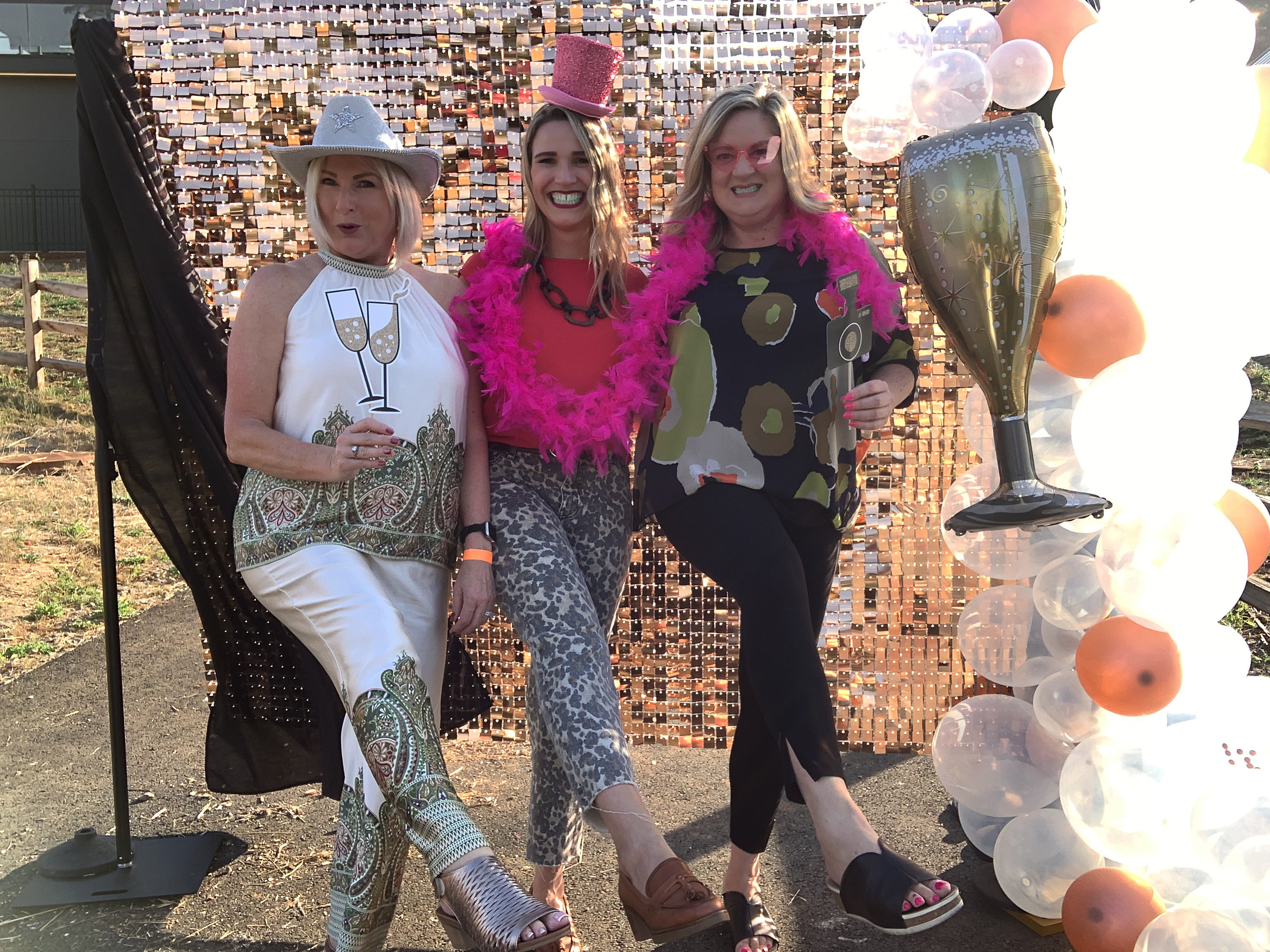Three friends posing at an outdoor party photo booth with a sequin backdrop, balloon arch, oversized champagne glass prop and pink feather boa.