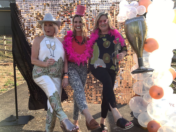 Three friends posing at an outdoor party photo booth with a sequin backdrop, balloon arch, oversized champagne glass prop and pink feather boa.