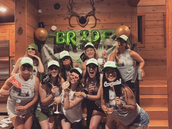 Smiling group of women at a rustic wood-paneled cabin bachelorette party, wearing matching green-and-white caps and tank tops, posing with BRIDE foil balloons, novelty photo props and drinks.