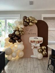 Neutral blush-and-chocolate arched party backdrop with gold, brown and cream balloon garland and oversized champagne coupe cutout in a bright living room celebration setup.