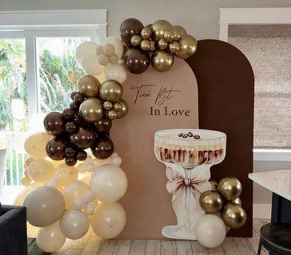 Neutral blush-and-chocolate arched party backdrop with gold, brown and cream balloon garland and oversized champagne coupe cutout in a bright living room celebration setup.