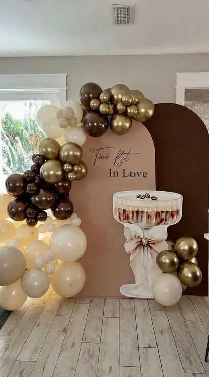 Neutral blush-and-chocolate arched party backdrop with gold, brown and cream balloon garland and oversized champagne coupe cutout in a bright living room celebration setup.