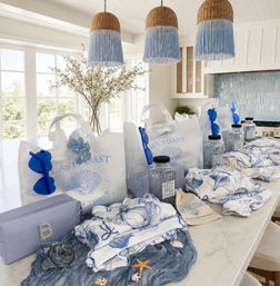 Coastal-themed blue and white gift bags and beachy accessories neatly arranged on a bright kitchen island — shell-printed totes, blue sunglasses, clear water bottles, patterned towels and seashell decorations under woven pendant lights