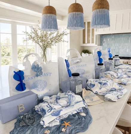 Coastal-themed blue and white gift bags and beachy accessories neatly arranged on a bright kitchen island — shell-printed totes, blue sunglasses, clear water bottles, patterned towels and seashell decorations under woven pendant lights