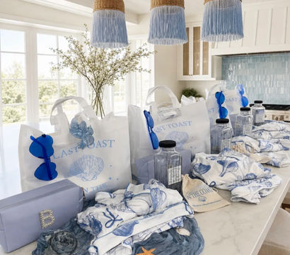 Coastal-themed blue and white gift bags and beachy accessories neatly arranged on a bright kitchen island — shell-printed totes, blue sunglasses, clear water bottles, patterned towels and seashell decorations under woven pendant lights