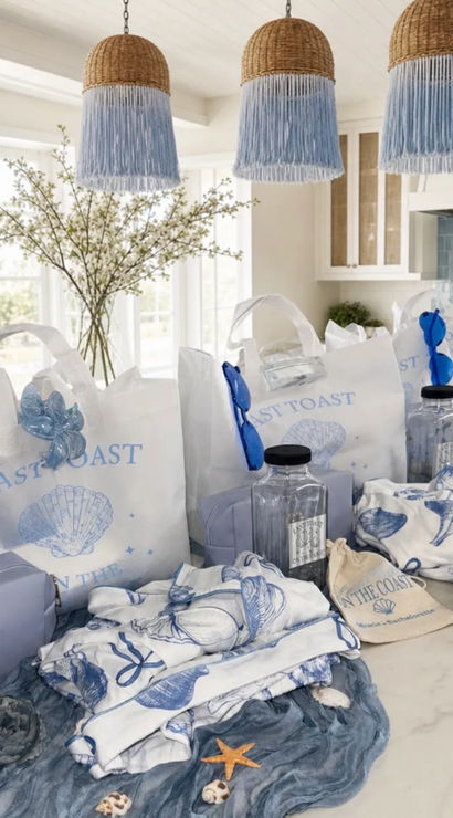 Coastal-themed blue and white gift bags and beachy accessories neatly arranged on a bright kitchen island — shell-printed totes, blue sunglasses, clear water bottles, patterned towels and seashell decorations under woven pendant lights