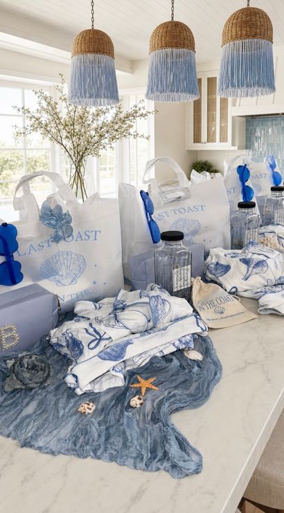 Coastal-themed blue and white gift bags and beachy accessories neatly arranged on a bright kitchen island — shell-printed totes, blue sunglasses, clear water bottles, patterned towels and seashell decorations under woven pendant lights