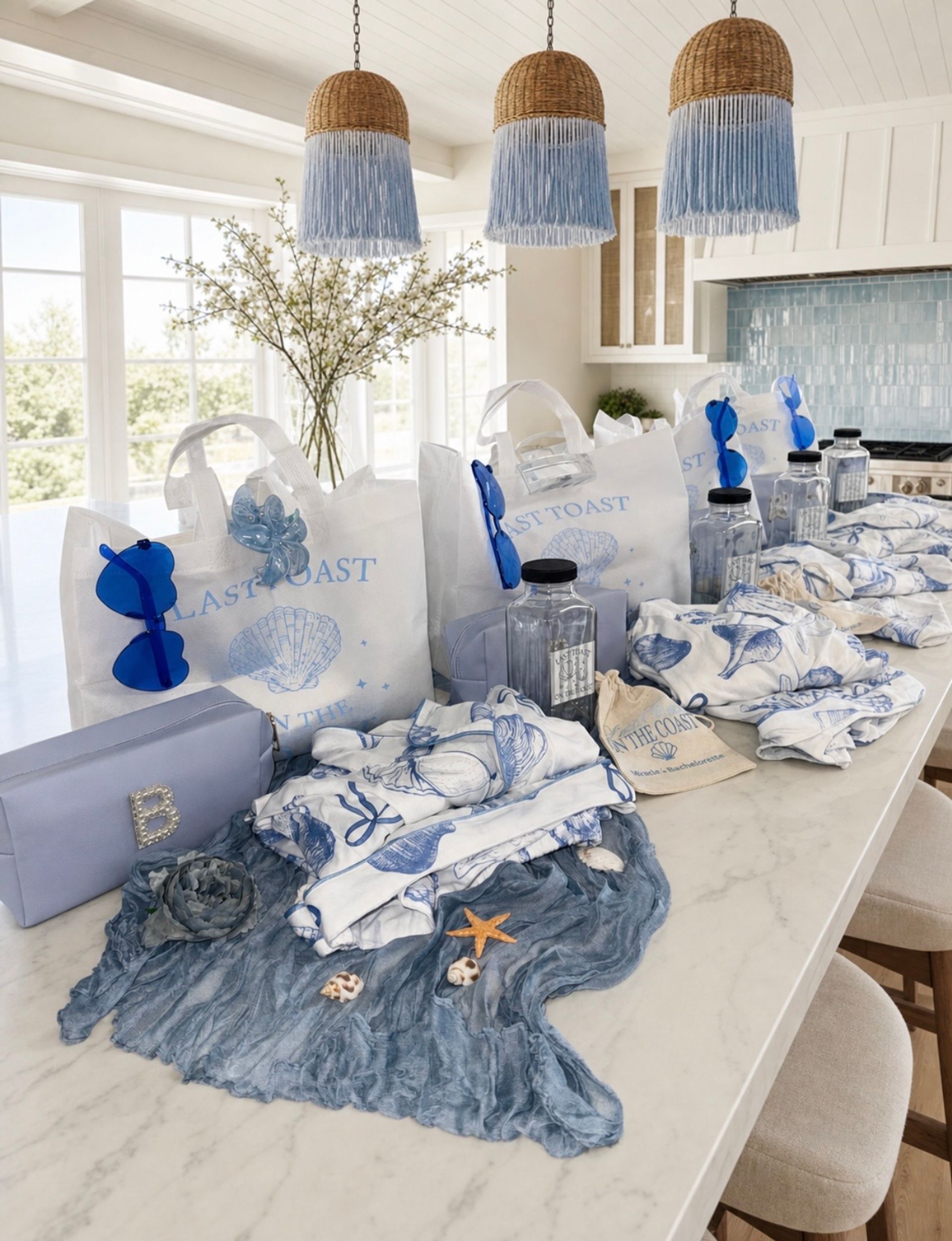 Coastal-themed blue and white gift bags and beachy accessories neatly arranged on a bright kitchen island — shell-printed totes, blue sunglasses, clear water bottles, patterned towels and seashell decorations under woven pendant lights