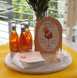 Spritz bar cocktail station with three orange aperitif spritz bottles, striped menu sign listing a spritz recipe, stacked napkins and glowing LED candles on a round marble board by a sunlit patio window.